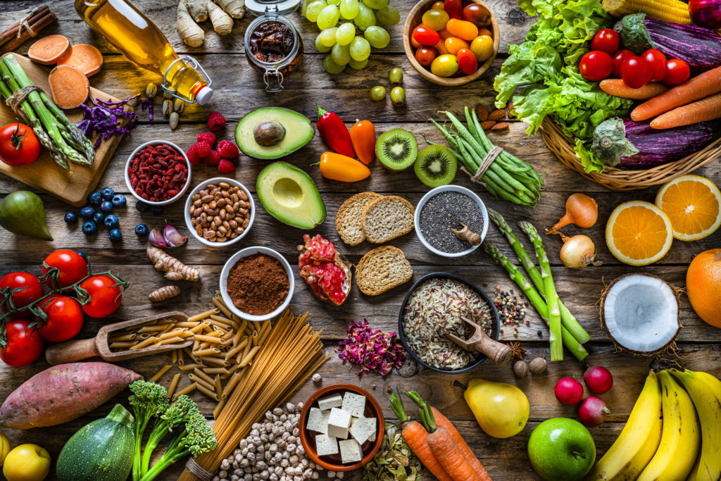 Vegan food backgrounds: large group of multicolored fresh fruits, vegetables, cereals and spices shot from above on wooden background. The composition includes green apple, kiwi, pear, pomegranate, orange, coconut, banana, grape, berries, ginger, almonds, pistachio, olive oil, olives, goji berries, chia seeds, pinto beans, nutmeg, rosemary, radish, tomatoes, carrot, kale, avocado, onion, rice, cocoa powder, sweet potato, wholegrain pasta, tofu, lettuce, corn, broccoli, pepper, asparagus, green beans, among others. High resolution 42Mp studio digital capture taken with SONY A7rII and Zeiss Batis 40mm F2.0 CF lens