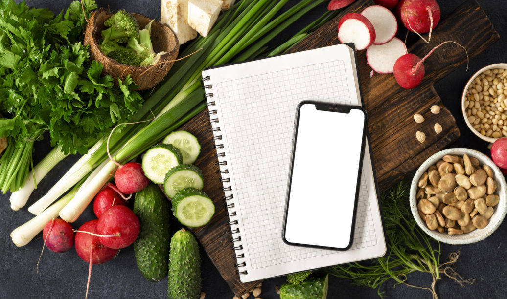 Order food online concept. Smartphone with blank screen and notepad with ingredients for cooking vegan food. Fresh vegetables, herbs, cereal and nuts on dark background top view